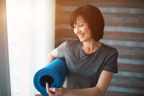 Adulte en forme femme mince a séance d'entraînement à la maison. Joyeux positif senior femme tiennent tapis de yoga dans la main et pointent sur elle. Sourire en restant debout dans la pièce avant l'entraînement . — Photo