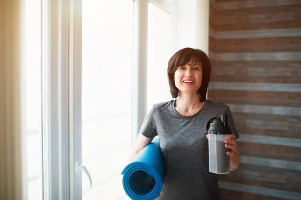 Vuxen passform smal kvinna har träning hemma. Positivt glad senior kvinna tittar på kameran och ler. Håller proteinshake och yogamatta i händerna efter träning. Vila och koppla av. — Stockfoto