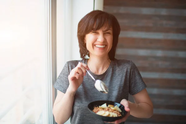 Vuxen passar smal kvinna som äter frukost ensam. Hålla skålen i handen och glad leende. Senior välbyggd person ta hand om sin hälsa. — Stockfoto