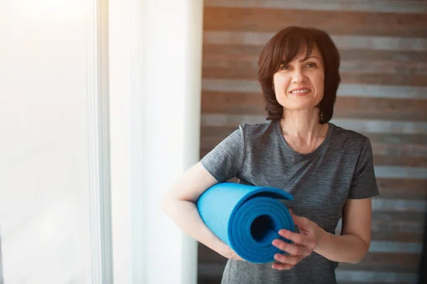 Vuxen passform smal kvinna har träning hemma. Välbyggd vuxen kvinna håller yogamattan i händerna och tittar på kameran. Ta hand om snygg och hälsa. — Stockfoto