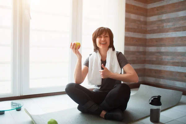 Vuxen passform smal kvinna har träning hemma. Sitt ensam på yogamattan med äpple i handen. Hälsosamt mellanmål efter träningen. Välbyggd stark kvinna på bild. — Stockfoto