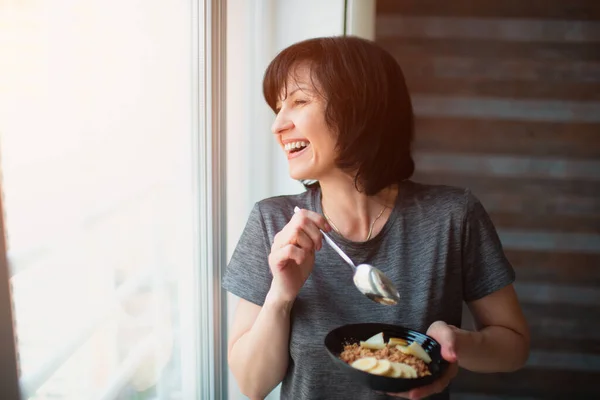 Vuxen passform smal kvinna äter frukost hemma. Ta hand om hälsa och välbefinnande. En kvinnlig pensionär som äter frukost i skålen. Titta på fönstret. — Stockfoto
