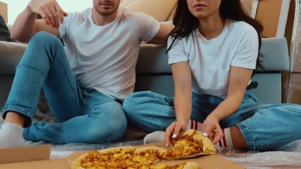 Una pareja joven se muda a un apartamento nuevo. Movimiento lento del hombre y la mujer sentados juntos en el suelo y comiendo pizza. Gente celebrando la reubicación en su nuevo apartamento . — Vídeos de Stock