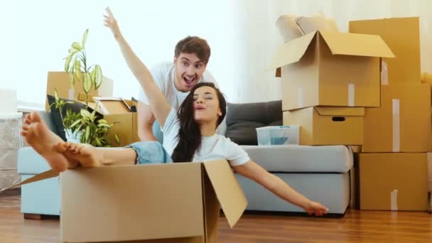 Młoda para wprowadza się do nowego mieszkania. Powolny ruch faceta pchającego pudełko ze swoją dziewczyną na podłodze i bawiącego się razem. Dziewczyny krzyczą i radują się. — Wideo stockowe