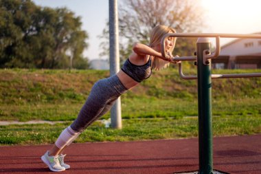 Kadın spor salonunda çalışıyor. Dışarıdaki ekipmanlarla şınav çekiyor. Eğleniyor..