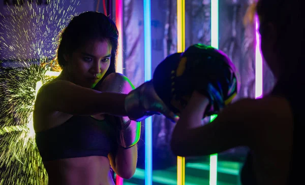 Beautiful 20s Asian Woman trains punching with coach on Silver gold Mitts Gloves. Office Girl exercise at Modern multi color Fashion Neon Muay Thai Boxing Gym with sweat water splash reflect light