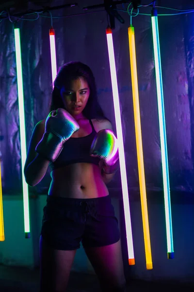 Beautiful 20s Asian Woman punch, poses with Silver gold Mitts Gloves. Office Girl exercise between Modern multi color Fashion Neon Muay Thai Boxing Gym with sweat water splash