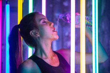 20 'li yaşlardaki güzel Asyalı kadın spor sutyen eğitimini tamamladı. Ofis kızı, modern çok renkli Neon Muay Tai Boks Salonu 'nda terli su sıçratarak temiz şişeden ağıza su içiyor.