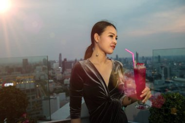 Gece elbisesi giymiş Asyalı kadın otelin çatısında dikiliyordu. Şehir manzarasında soğuk kokteyl kırmızı meyve suyu ve alacakaranlık periyodu vardı.