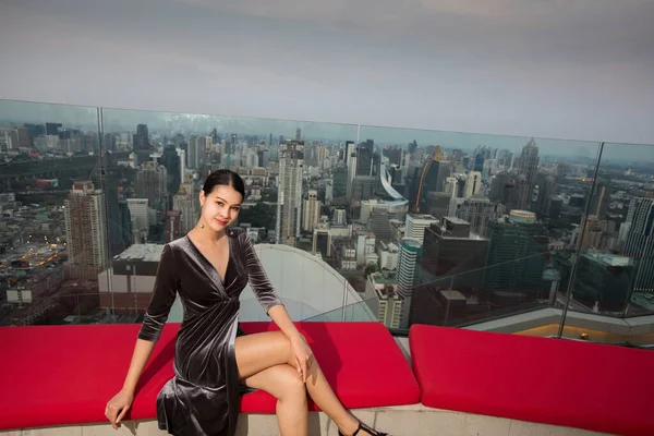 身着晚礼服的亚洲女人坐在城市景观城市酒店顶部的红色沙发上 有着水平的暮色 — 图库照片