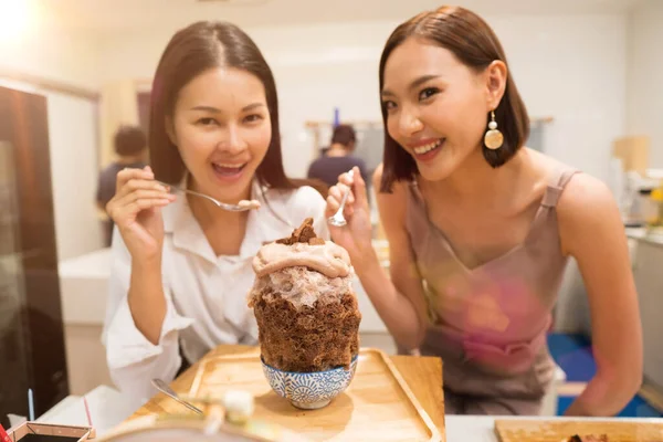 Two Beautiful Adult working women eat chocolate Bingsu ice with sweet juice dessert on top  atmosphere with many food plates and people in restaurant, selective focus on bingsu blur people