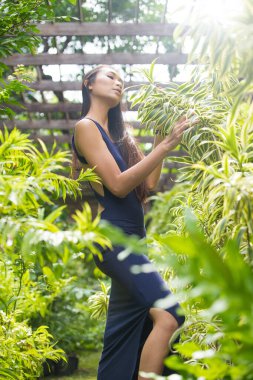 Güzel Asyalı düz düz siyah saçlı koyu mavi açık arka elbiseli kadın. İnce, fotokopi odası. Ormanın ortasında çeşitli yeşil bitki yaprakları var.
