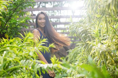 Güzel Asyalı düz düz siyah saçlı koyu mavi açık arka elbiseli kadın. İnce, fotokopi alanı. Ormanın ortasında çeşitli yeşil bitkiler var.