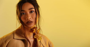 Güzel Asyalı Uzun düz siyah saçlı kadın ceket önlüğü, makyaj malzemesi, stüdyo ışıkları sarı arka plan izole edilmiş kopya alanı, altın çiçek