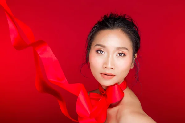 Fashion portrait of Asian Black hair tanned skin woman with strong color red lips, studio lighting red reddish background copy space, girl ties bow red ribbon and wind blow, selective focus