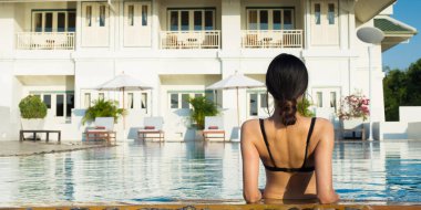 Bronzlaşmış tenli, ince Asyalı kadın siyah bikinili makyaj gözlüklü, güneş gözlüklü, kocaman mavi yüzme havuzu boyunca gülücük yollayan, güzel gökyüzü tatil tarzı, metin logosu için kopya alan.