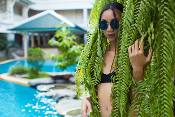 Beautiful Tanned skin Asian slim woman in black lace bikini swimwear fashion make up, Post play strong in green leaf tree along big blue pool during vacation holiday, copy space for text logo