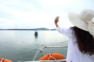 Asya Yarışı bronz tenli kadın Bikini Beyaz Gömlekli Büyük Şapkalı kısa kot pantolon Yat teknesinin güvertesinde turuncu can simidi ile yaz tatilinde mavi gökyüzü altında, uzay logosunun kopyası.