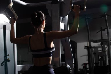 Asyalı zayıf fitness kadınının arkadan görünüşü siyah saçlı spor sutyen itme makinesinde ısınma, ağırlık çekme, kas gücü, kollar çok işlevli spor salonunda. Kadın sporcu sağlıklı sporcu olabilir.