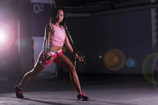 Tan Skin Woman in Fitness pink Suit red short do heavy TRX rope push up exercise training at crossfit Garage, dark shadow studio lighting, look left, copy space for text logo