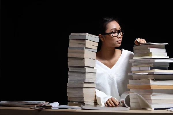 Beautiful Asian Girl open and read magazine between stacking of many kind books, textbooks, journal, report, research, comic in two towers on table with dark black background night time, copy space