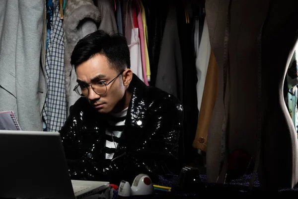 Black Sequin jacket of Asian Fashion Designer Man checks order and sale growth on Notebook computer. Gay tailor designs new trend of collection to Cloth rack backgrounds, low key exposure copy space