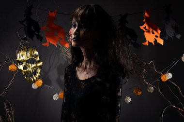 Cadılar Bayramı geçmişinde cadı ve balkabağı çelengi asılı, Yüzünde taze yara olan hayalet zombi kız Yüzünde siyah saç elbisesi korkunç ve tüm perili yerleri öldürmek istiyor