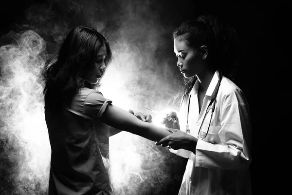 Asian Beautiful Doctor Nurse woman in medical uniform with stethoscope inject vitamin syringe to patient to treat healthcare, studio lighting dark smoke backgrounds copy space, black white monochrome