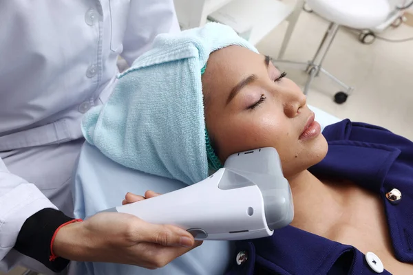 Asyalı kadına, cildini pürüzsüz kılmak ve sivilceleri tedavi etmek için hastane kliniğindeki soğuk algınlığı aleti ile tedavi etmek için sabırlı davranılmalıdır.