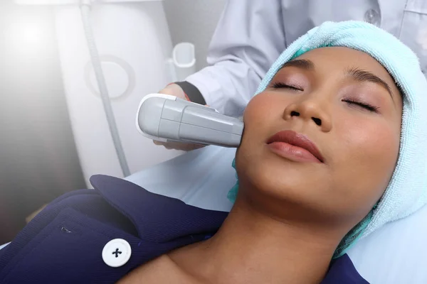 Asyalı kadına, cildini pürüzsüz kılmak ve sivilceleri tedavi etmek için hastane kliniğindeki soğuk algınlığı aleti ile tedavi etmek için sabırlı davranılmalıdır.