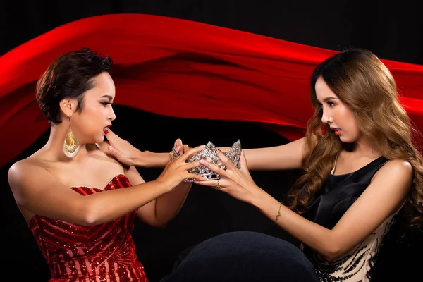 Two Miss Beauty Pageant Queen Contestants fight, catfight, for a jewel Diamond Crown. They wear Sequin Long Gown and struggle for the prize in the background of red cloth flying in the Air