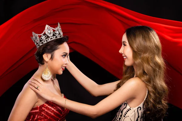 Two Miss Beauty Pageant Queen Contestants happy and smile for a jewel Diamond Crown. They wear Sequin Long Gown and give put the prize on Head in the background of red cloth flying in the Air