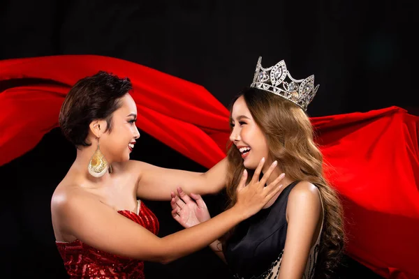 Two Miss Beauty Pageant Queen Contestants happy and smile for a jewel Diamond Crown. They wear Sequin Long Gown and give put the prize on Head in the background of red cloth flying in the Air