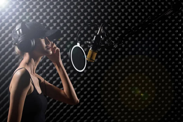 ミックスレーススキニー女性ブロンドの髪キャップイヤーマフヘッドフォンは ハングマイクコンデンサーの上にパワーサウンドで大声で歌を歌います 卵クレートスタジオ低照明影防音壁の部屋を吸収 — ストック写真