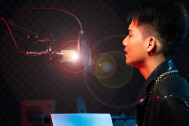 Asyalı genç adam siyah saç teli kulaklığı mikrofon kondansatörü, hoparlörler, besteci not defteri ve monitör üzerinde yüksek sesle bir şarkı söylüyor. Stüdyo Ses Algılama Duvarı Odası