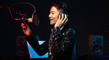 Asyalı genç adam siyah saç teli kulaklığı mikrofon kondansatörü, hoparlörler, besteci not defteri ve monitör üzerinde yüksek sesle bir şarkı söylüyor. Stüdyo Ses Algılama Duvarı Odası