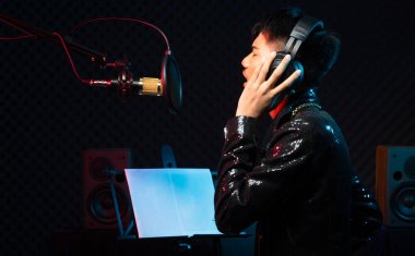 Asyalı genç adam siyah saç teli kulaklığı mikrofon kondansatörü, hoparlörler, besteci not defteri ve monitör üzerinde yüksek sesle bir şarkı söylüyor. Stüdyo Ses Algılama Duvarı Odası