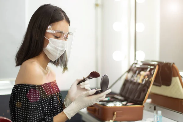 Covid New Normal of Career working with people, make up artist has to wear rubber glove, surgical mask and Face Shield to protect both herself and customer. Young Asian Woman hold brush powder