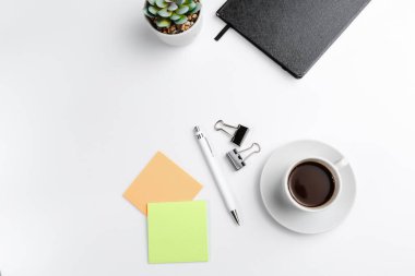 Siyah, kapalı bir Not Defteri, kahve fincanı, renkli ofis etiketleri ve beyaz arka planda çiçek manzaralı bir kalem. Metin için boşluk