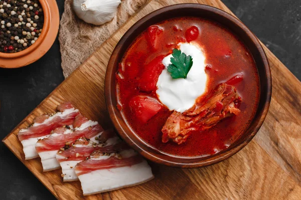 Ukrayna Borsch 'u. Pancar çorbası. Ukrayna mutfağı. Ahşap bir tabakta tuzlanmış pastırma, ekmek ve otlarla birlikte. Karanlık arka planda. Yukarıdan manzara. Metnin için boş yer. Bir kase kremalı kırmızı pancar çorbası..