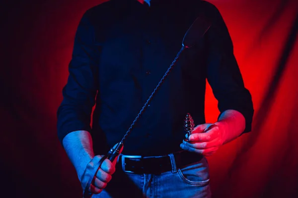 Bir adam elinde kırbaç ve elinde anal tıkaçla kırmızı bir arka planda duruyor. — Stok fotoğraf