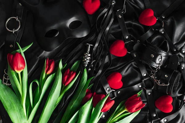 Explore the world of kinky play. Top view of bdsm leather kit against of a black silk. tulips