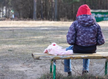 Gri ceketli üzgün bir çocuk, yanında bir oyuncakla bankta tek başına oturuyor.