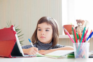 Kız öğrenci 8 yaşında uzaktan öğrenim görüyor. Tabletle dersler alıyor. Masada oturan bir not defterine yazıyor.