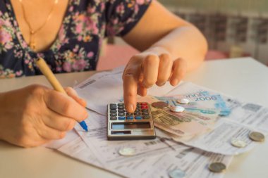 Yaşlı bir kadın faturalar için ödeme yapmayı düşünür; bir hesap makinesinde, Rus rublesi içinde parası olan bir daire için ödeme yapmayı düşünür; 2000,100 ruble ve Rus sikkesi banknotları