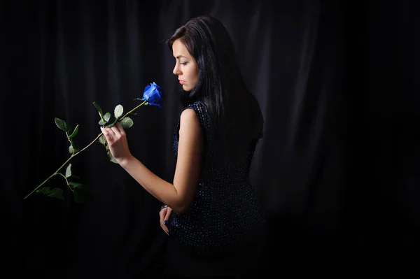 Girl with shadowy hair in a black dress looks at a blue rose, low key