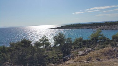 Denizin üzerindeki kayalık yamaç, yamacın üstündeki ağaçlar, mavi deniz.