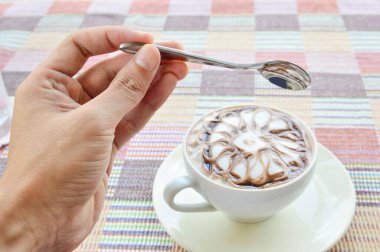 Elinde bir kaşık, çiçekli bir fincan kahve renkli kumaş arka planda beyaz bir fincanda.