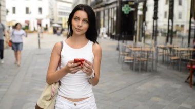 Beyaz elbiseli genç siyah kadın sakin caddede yürüyor, etrafa bakıyor ve Bratislava 'nın tarihi bölgesinde akıllı telefon navigasyonunu kullanıyor. HD VIDEO