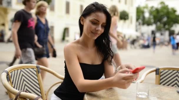 ブラチスラバ市の歴史的な地域にあるコーヒーバーのテラスのテーブルで携帯電話で遊んでいる美しい笑顔の女の子 Hdビデオ — ストック動画